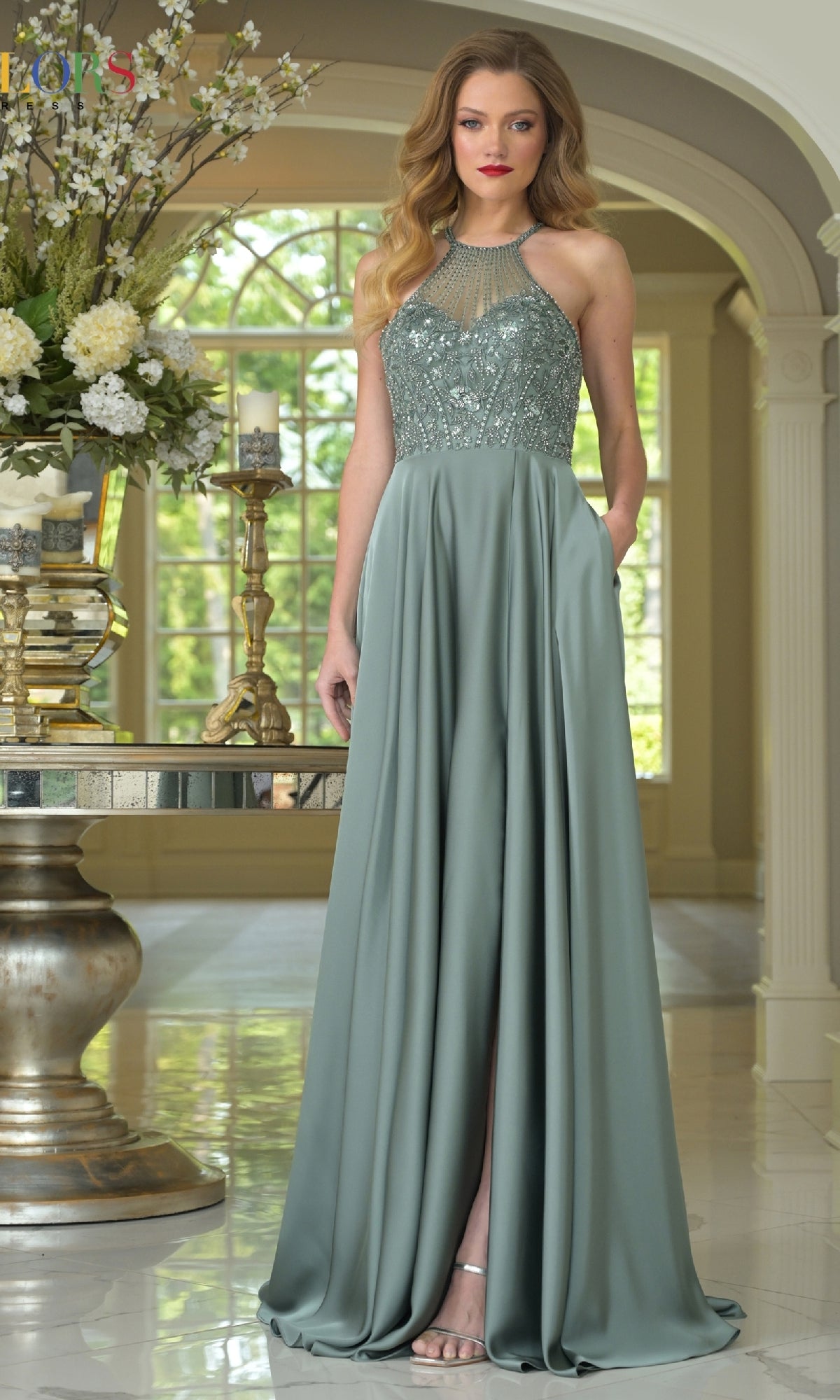 A woman wears the Colors Dress High-Neck Halter Prom Gown (Style 3822) in sage green, featuring a beaded bodice and flowing skirt, standing indoors by a mirrored table with flowers, candles, and a gold vase near large windows.